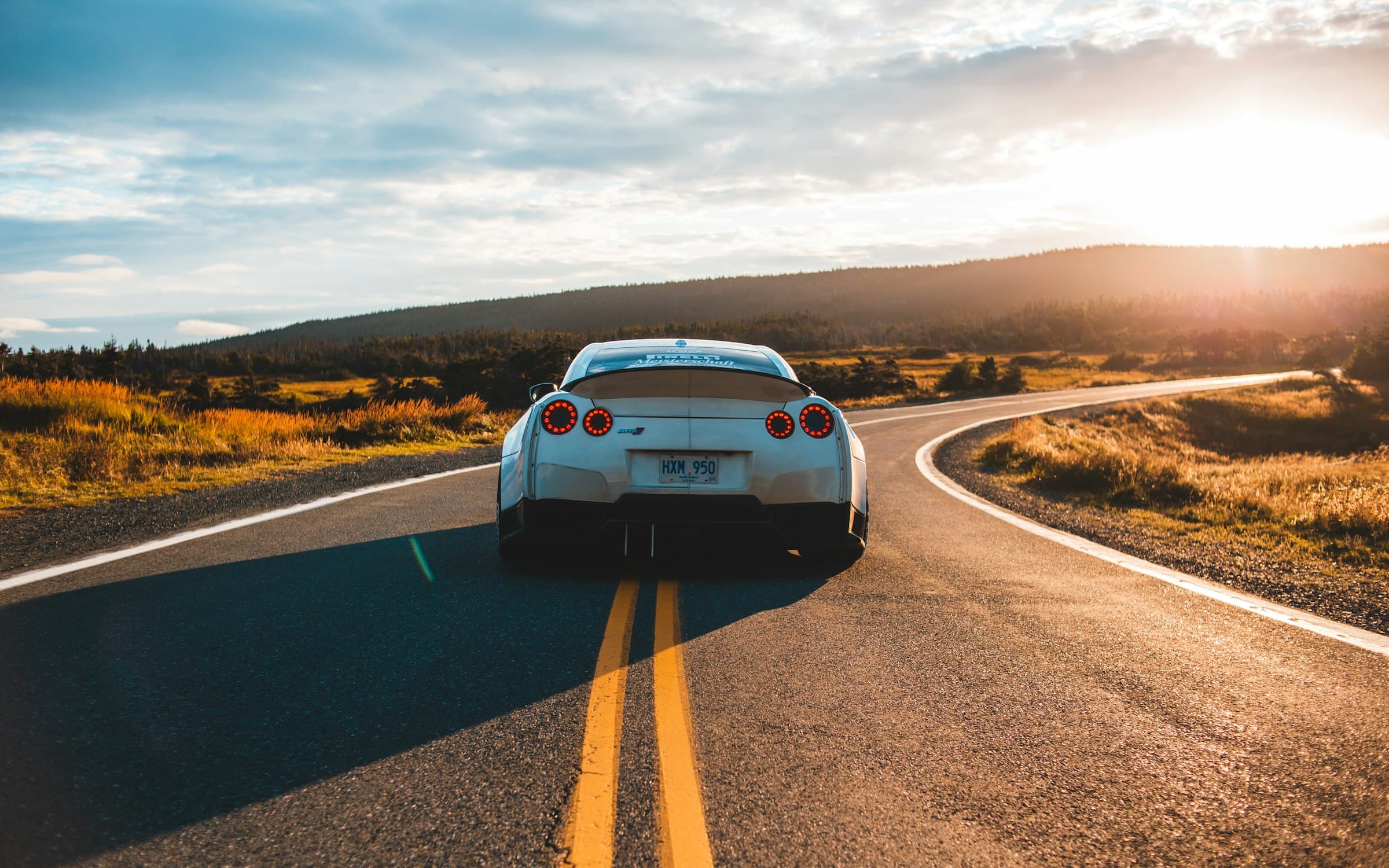 A white sports car driving away down a winding two-lane road at sunset
